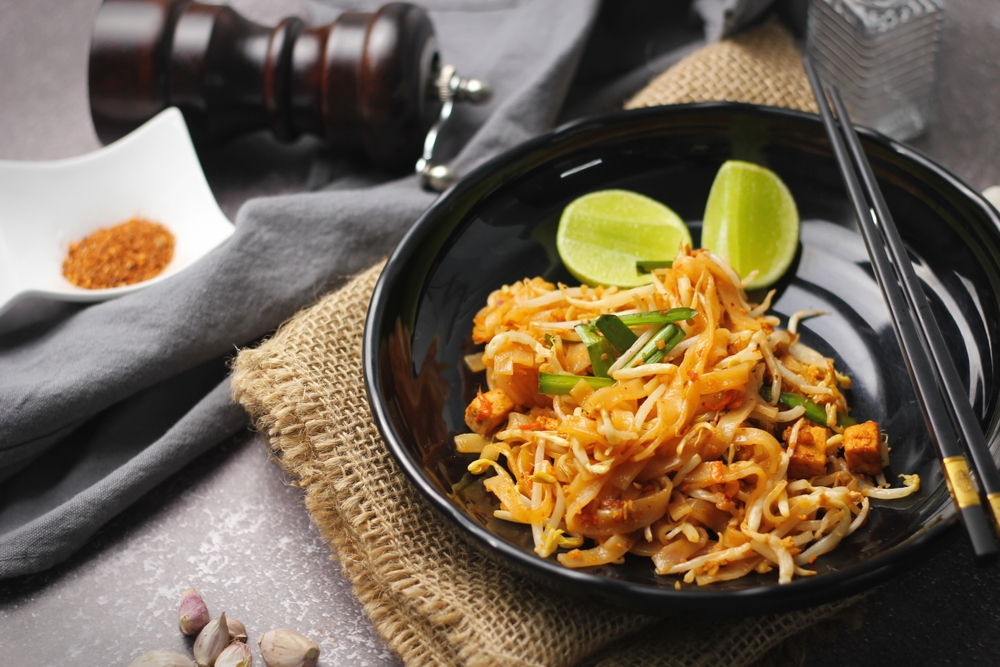 A black bowl of pad thai noodles garnished with lime wedges and green onions, set on a burlap cloth with chopsticks. Nearby are garlic cloves, pepper mill, and spice.