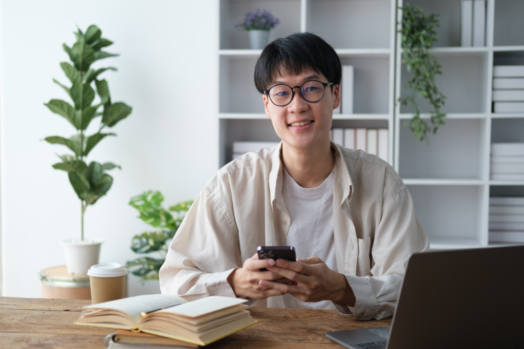 Man sitting at a desk uses his phone with a laptop, notebook, and coffee nearby