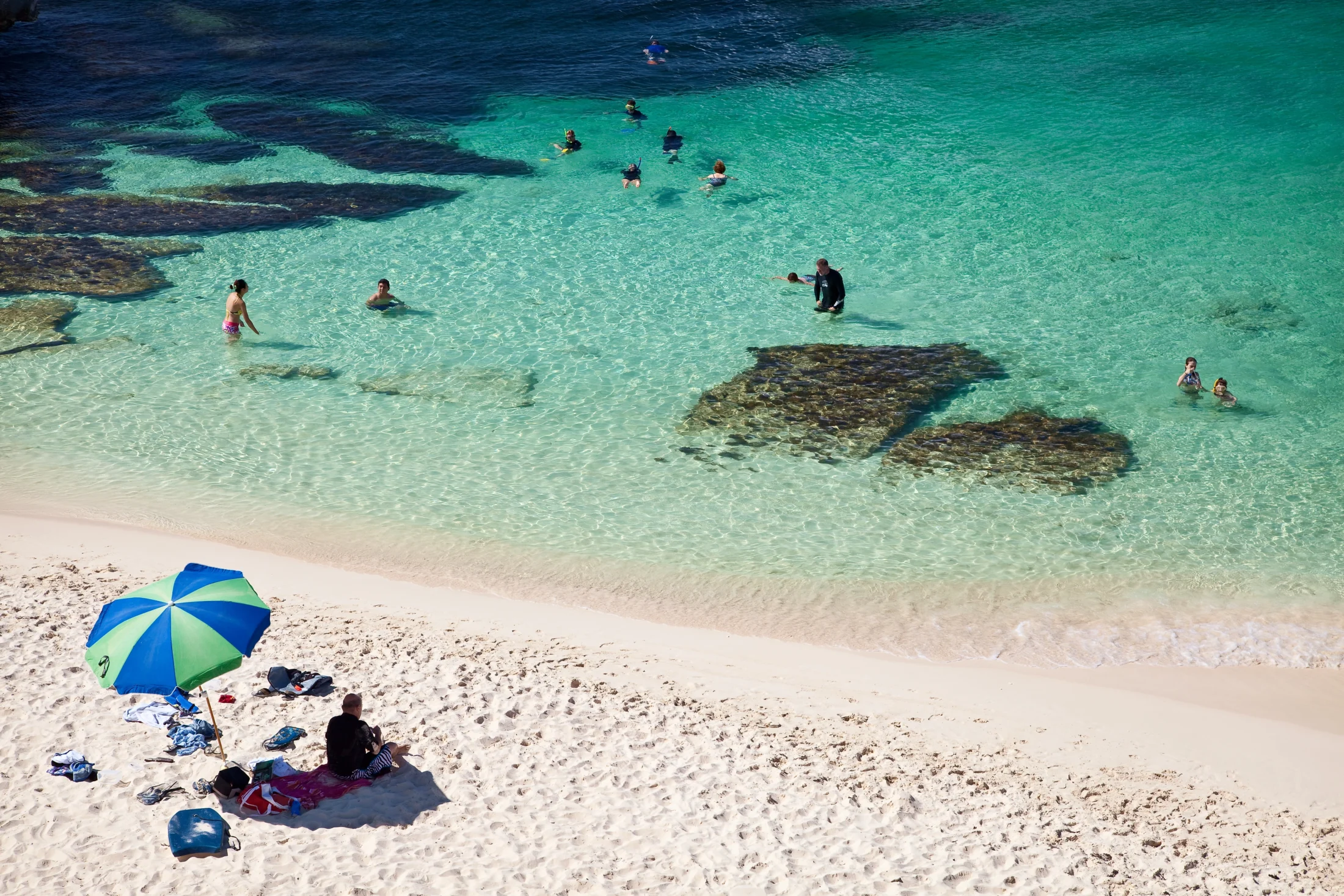 Sandy beach on Rottnest Island, Western Australia, with two people sitting under a green and blue umbrella in the foreground. Clear turquoise water stretches to the horizon, where several people are swimming near darker rock formations beneath the surface.