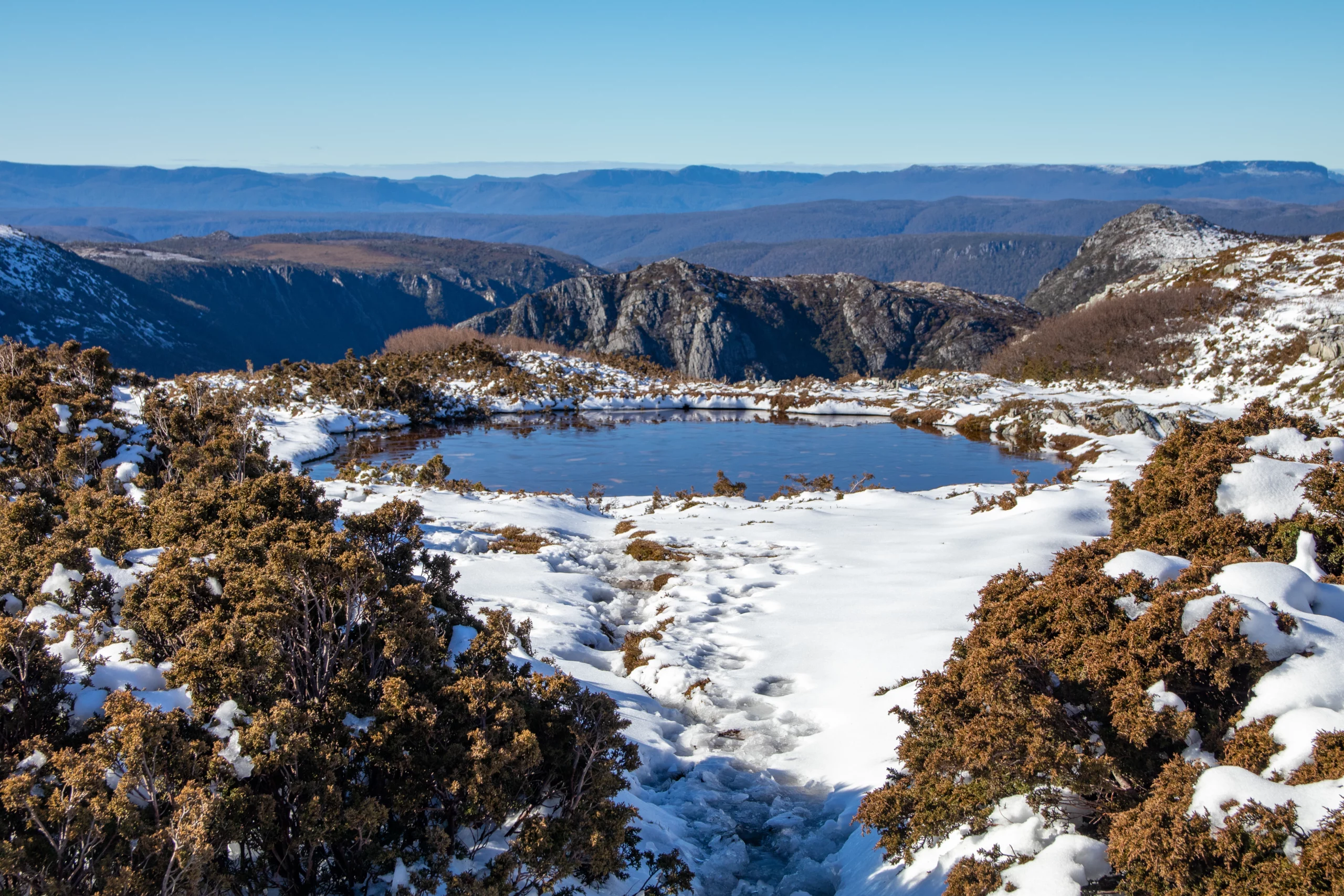 Snow-covered alpine landscape in Cradle Mountain-Lake St Clair National Park, Tasmania, featuring a partially frozen mountain lake surrounded by rocky peaks and low shrubs.