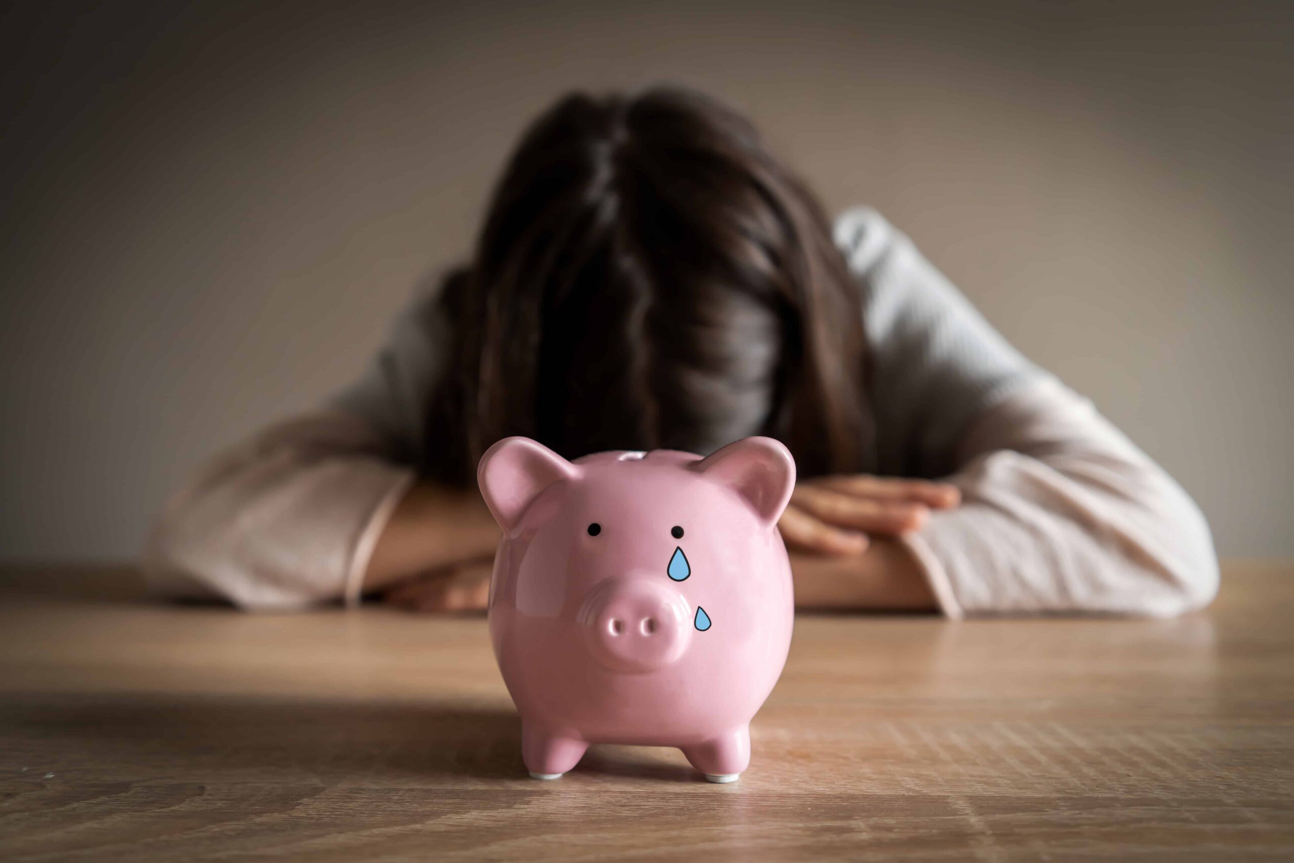 A pink piggy bank with a tear drop decal sits on a table. Behind it, a person with their head down appears dejected. The scene conveys sadness.