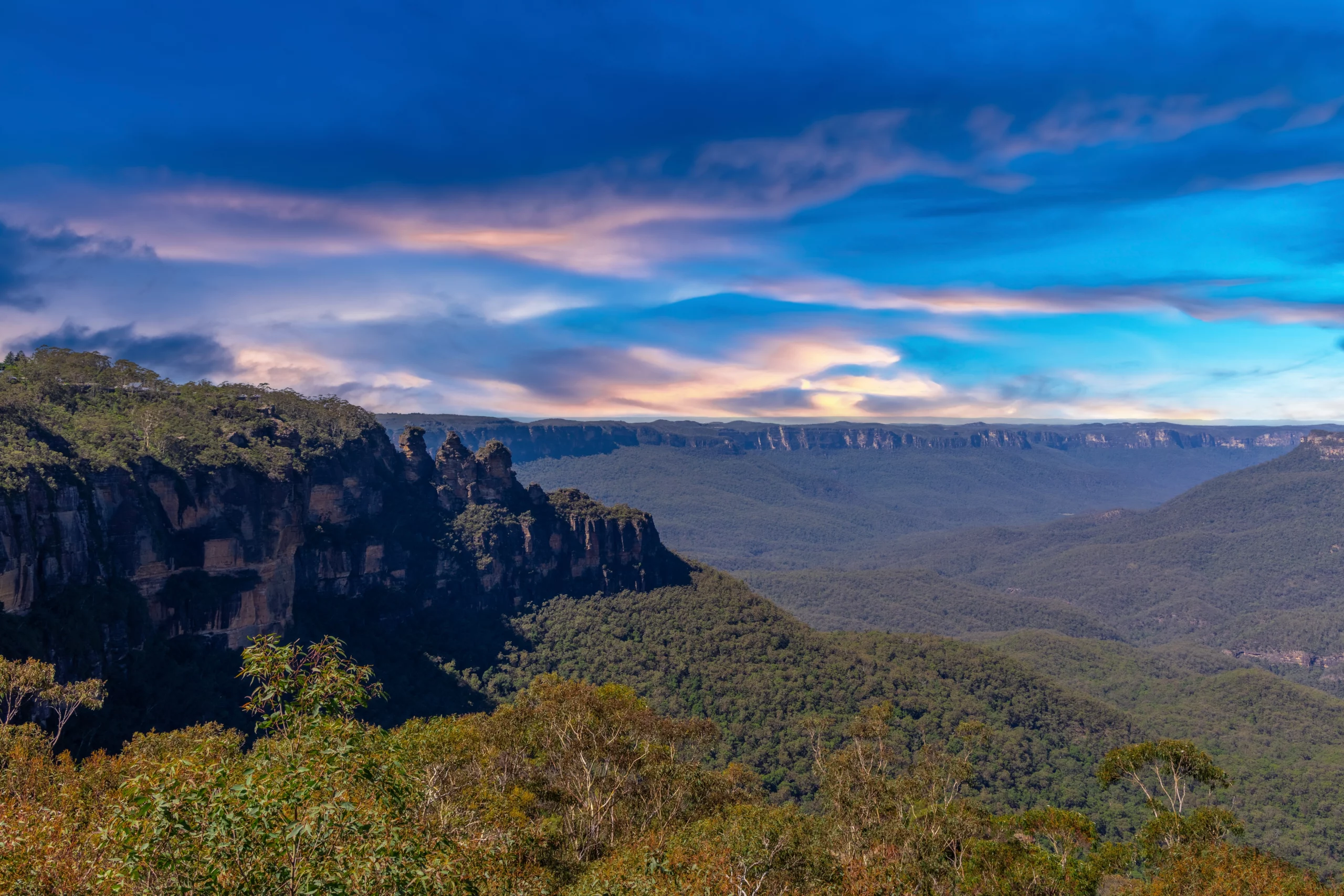 Expansive view across Blue Mountains National Park in New South Wales, with rugged sandstone cliffs, dense eucalyptus forest, and dramatic blue-toned skies stretching into the distance.