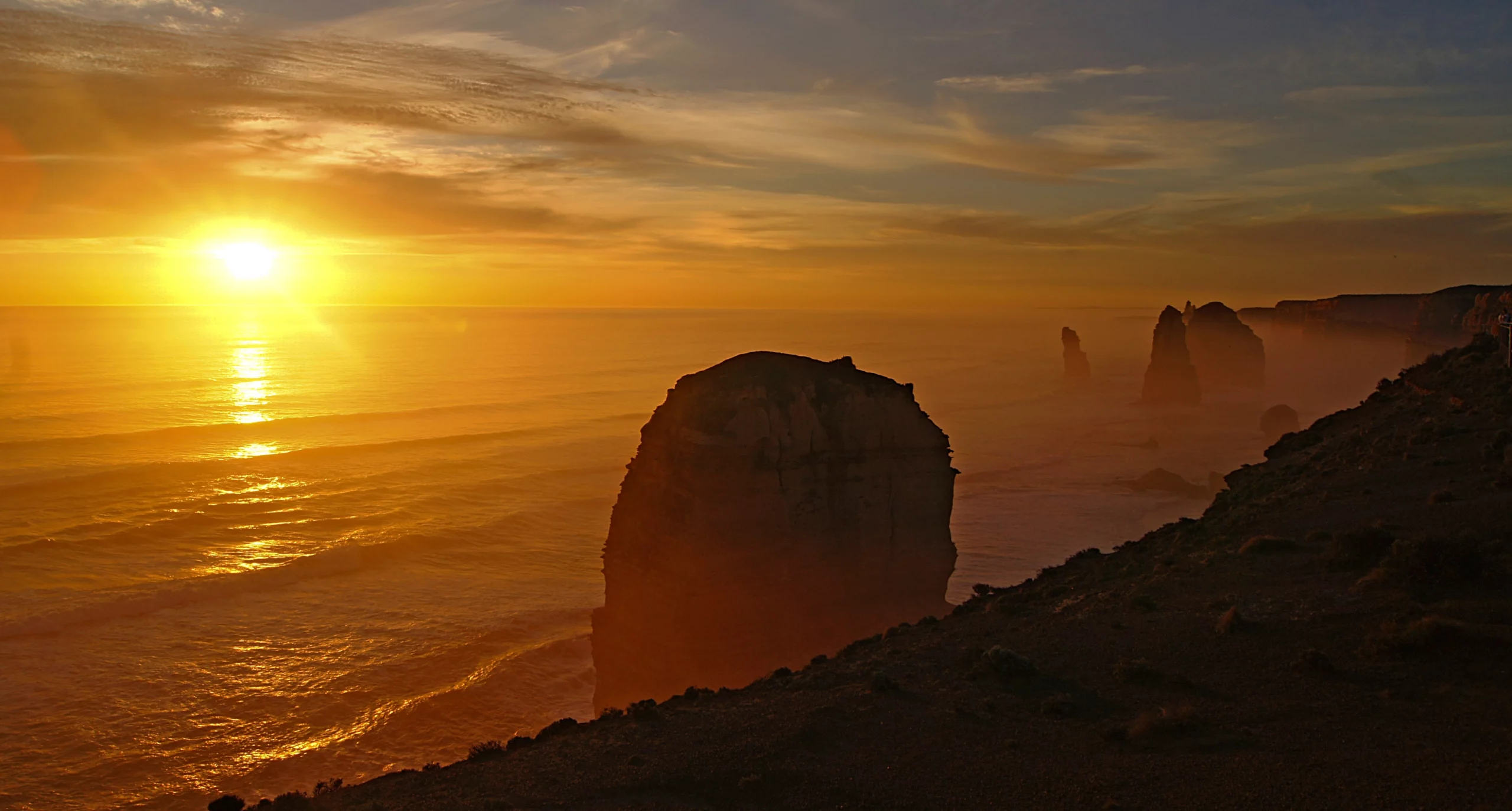 Sunset view along the Great Ocean Road in Victoria, featuring towering coastal rock formations rising from the ocean beneath an orange and golden sky.