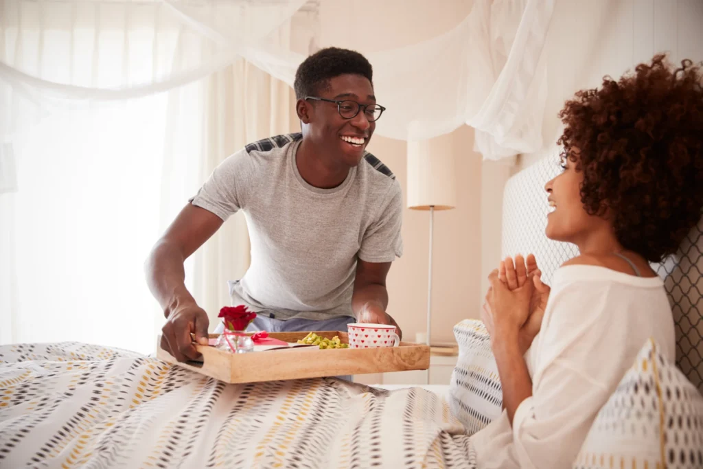 Father giving wife who is a mother a tray of food for breakfast in bed with a rose and heart-covered mug as a last minute Mother's Day gift