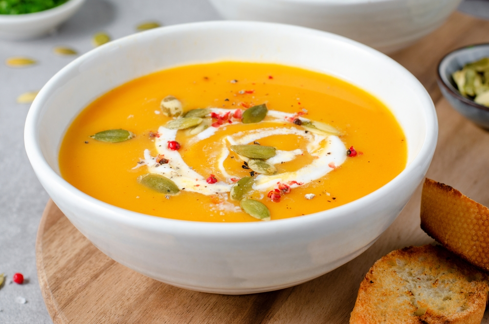 A bowl of creamy orange soup garnished with a swirl of cream, pumpkin seeds, and red pepper, served with toasted bread on a wooden board—a cheap easy lunch idea for work.