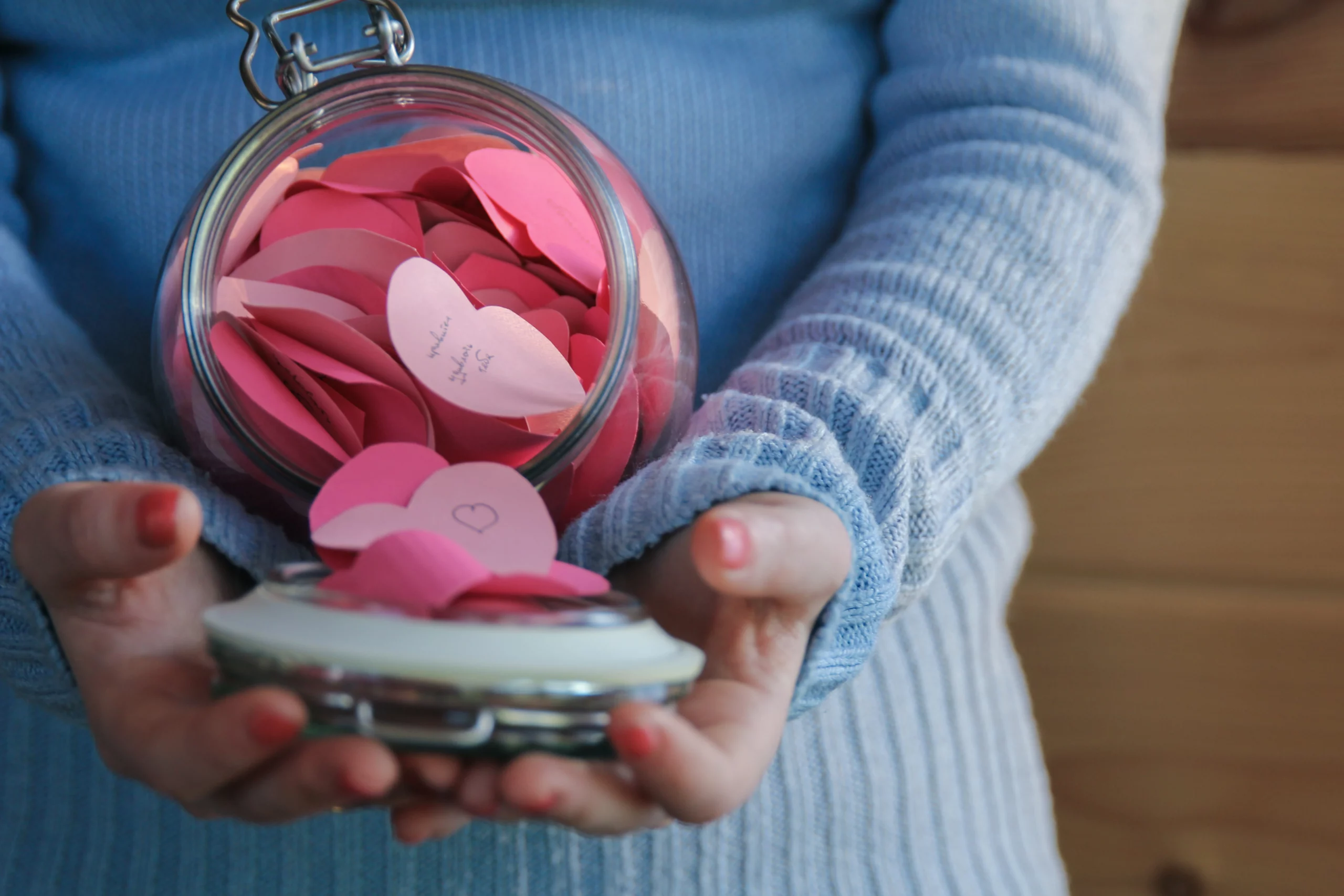 a woman holding a glass thankfulness jar filled with different shades of pink paper hearts with notes on them for a quick last minute Mother's Day gift