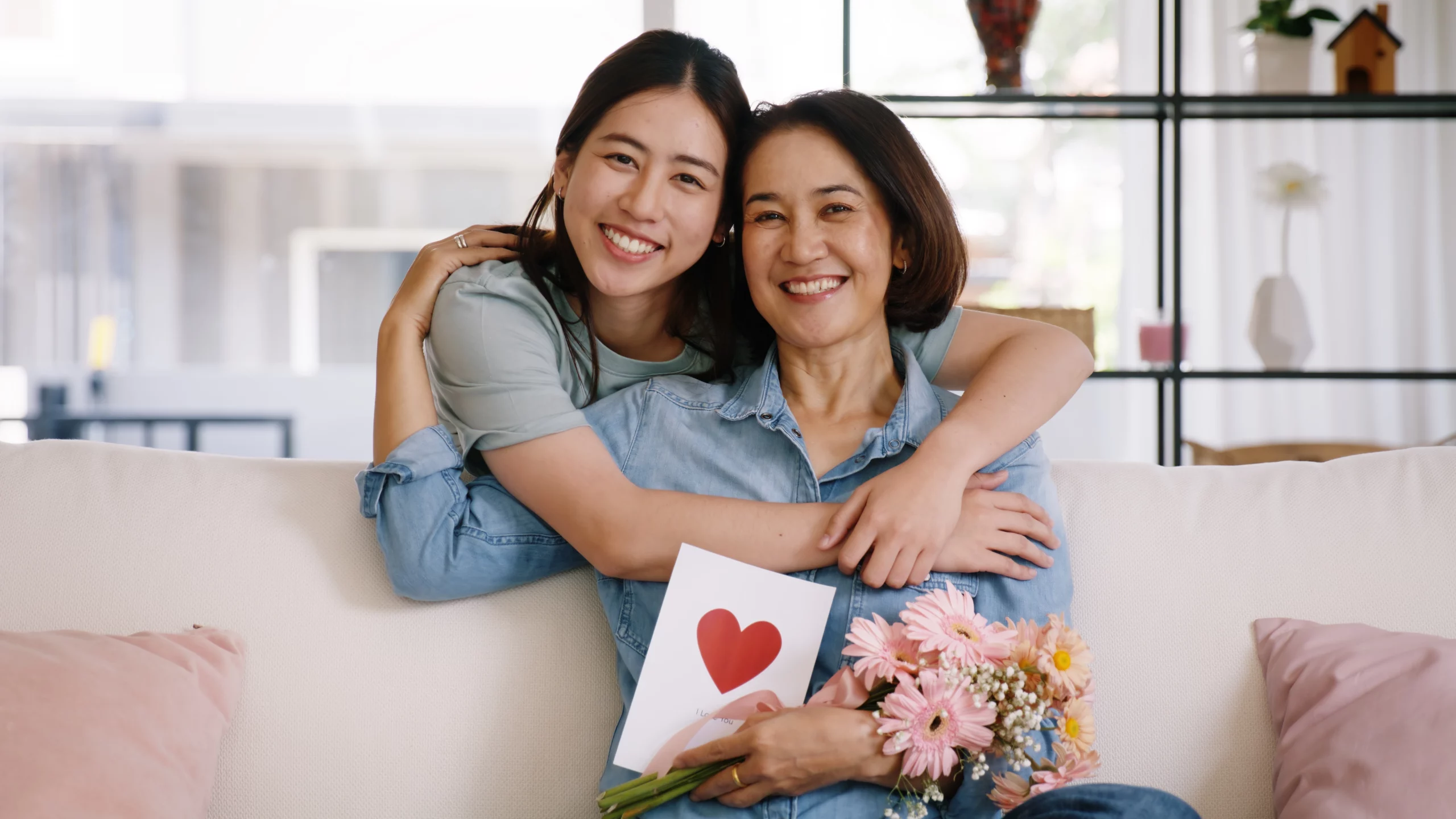 mother and daughter hugging on couch, with mother holding a handwritten card and bouquet given as a last minute Mother's Day gift