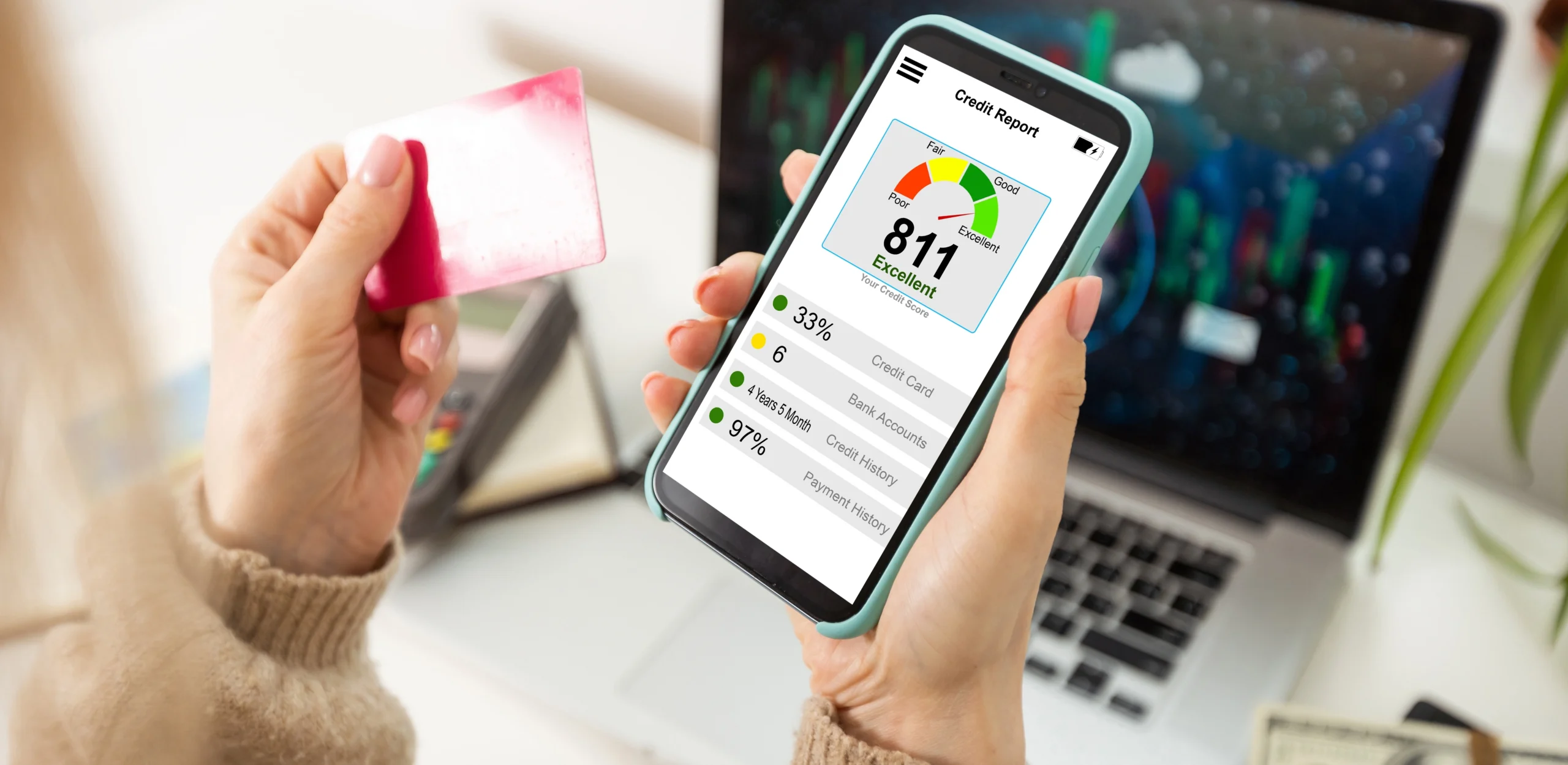 Close up of a person looking at their excellent credit score on their phone, holding a credit card in their other hand in front of a laptop