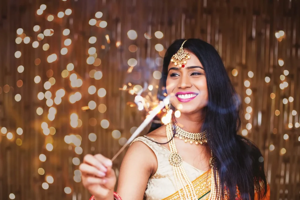 A smiling woman dressed in traditional Indian attire holds a sparkler during a festive celebration, with warm golden lights glowing in the background.