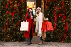 Two women wearing winter coats smile and hold shopping bags in front of a festive Christmas-themed storefront.