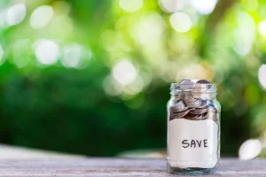 A jar filled with coins labeled "Save" sits on a wooden table. The blurred green foliage in the background conveys a peaceful, hopeful mood.