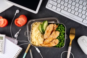 Lunchbox with grilled chicken, peas, cucumber, and couscous on a desk with a keyboard, mouse, and note pad, conveying a balanced work lunch.