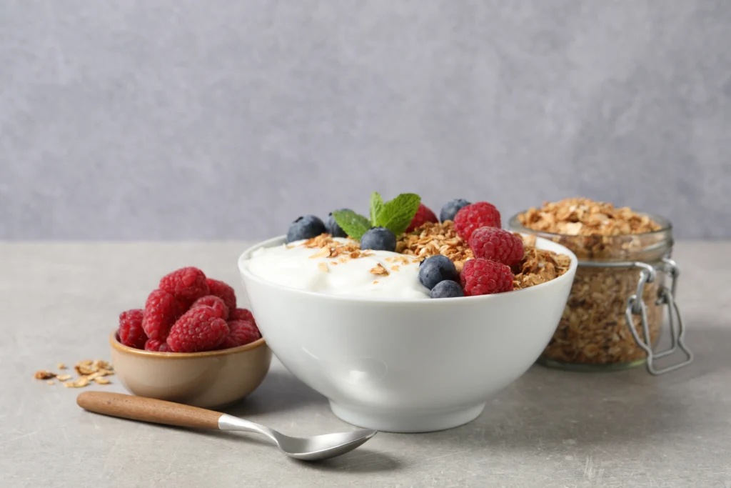 A white bowl of Greek yogurt topped with granola, raspberries, blueberries, and mint, accompanied by a smaller bowl of raspberries, a jar of granola, and a spoon.