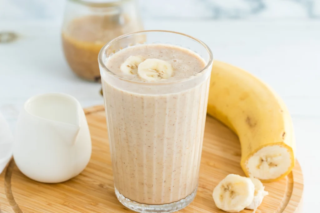 A creamy protein smoothie in a glass on a wooden tray, garnished with banana slices. Nearby are a whole banana and a small white pitcher, creating a fresh, inviting scene.