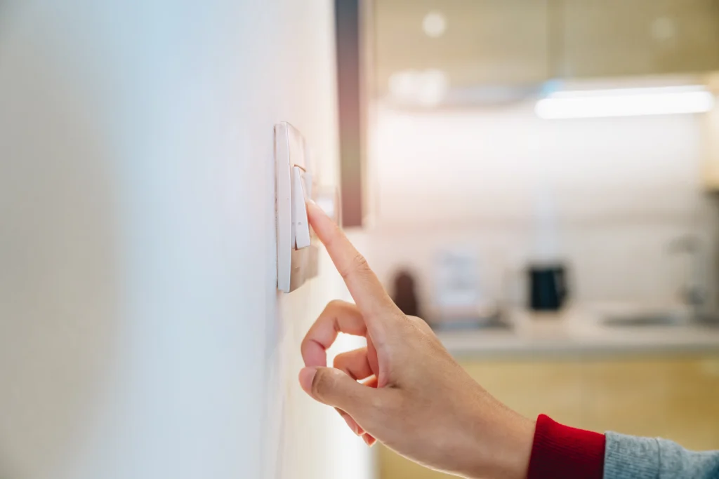 A hand presses a light switch on a white wall in a softly lit room, conveying calm and routine. Blurred kitchen elements are visible in the background.