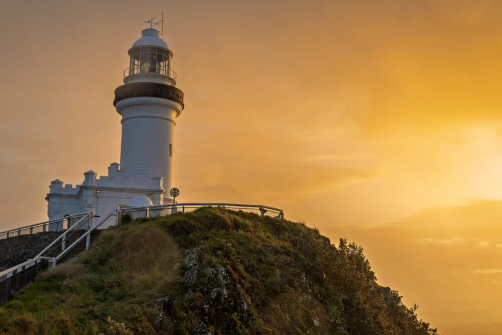 Cape Byron Lighthouse stands on a grassy, rocky hill under a golden sunset sky. The white cylindrical tower with its lantern room is framed by a fence and surrounding greenery.