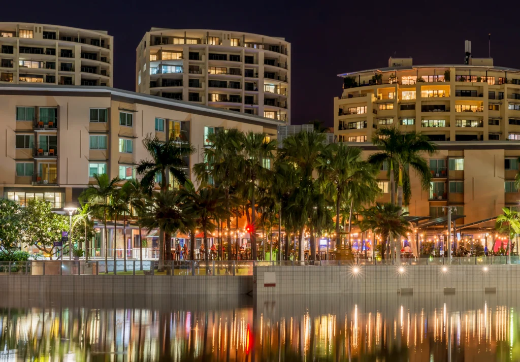Night view of the Darwin Waterfront with palm trees, lit buildings, and reflections on water.